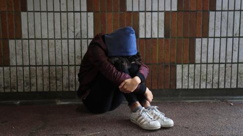 A hooded teenage girl sits in front of a tiled wall in pedestrian underpass  with her head resting on her knees