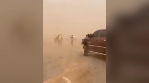 A maroon car sits on a dust-covered road immersed in a dust-cloud as people in high-vis jackets stand beyond it.