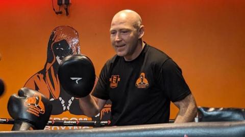 A boxing coach in a ring working with a young person in gloves, with their back to the camera