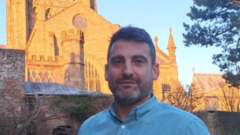 Sam Pratley standing outside Hereford Cathedral. It's a sunny day with a clear blue sky and Mr Pratley is wearing a blue and white striped shirt.