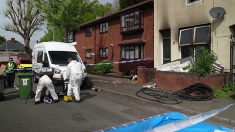The fire-hit property is on the right of the image with black smoke marks going up the front of the home above windows. Police tape is in place either side of the terraced home and forensic officers are within that in the street in white suits. Police can be seen on the other side of a the tape and a large white van is parked nearby by the pavement. 
