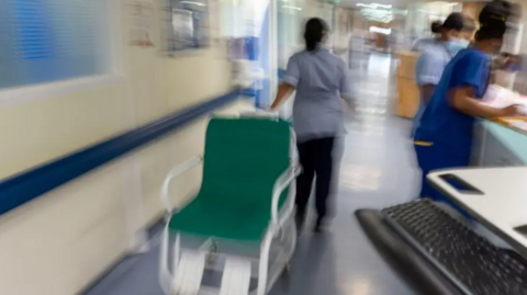 A nurse wearing a blue top pulling a green and white wheelchair down a hospital corridor. There are two other nurses on the right looking at paperwork. The image is blurred to show the movement of the nurse pulling the chair. The walls are white and has a blue border running through the middle.