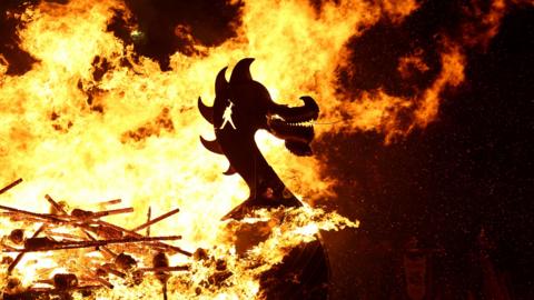 Flames rise around the carved head of a Viking long boat replica. Flaming torches have been thrown into the boat