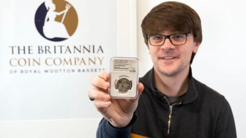 Christopher Collects, wearing black top and glasses, holding the coin in front of sign saying 'The Britannia Coin Company'.