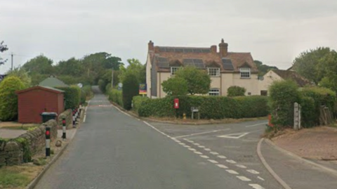 A long straight narrow village road on the left with a driveway and shed, and bushes and hedgerows along the side of it. On the right here is a small junction that heads past a cream house and green bushes.
