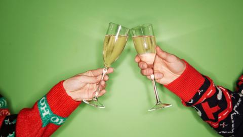 Two people wear Christmas jumpers and cheers with a glass of prosecco against a green background.
