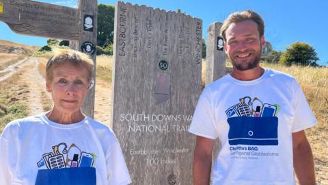 Alex and Miles standing by a sign which says Eastbourne to Winchester 100 miles. Both are wearing t-shirts for their charity.
