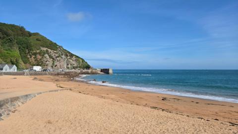 La Grève de Lecq in Jersey on a sunny day. Waves are lapping on the sandy shoreline. The sky is a vibrant blue. A green cliff leads down to the beach. 