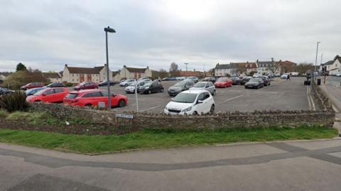 Rock Street Car Park in Thornbury. It is situated on a hill, on a bend in the road. It is surrounded by a brick wall and a pavement.