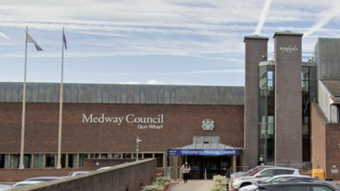 a long brown and grey brick building with Medway Council Gun Wharf written on the side of the building. A covered entrance has a blue and white sign saying "Welcome to Medway Council". A woman is seen walking from the entrance towards the car park outside where a line of cars are parked.