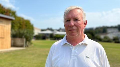 A head-and-shoulders shot of a man with grey hair, wearing a white polo shirt with a small green crocodile logo. He is standing outdoors at millennium park's  grassy area. In the background, there is a small wooden building, as well as other buildings and trees under a cloudy sky.
