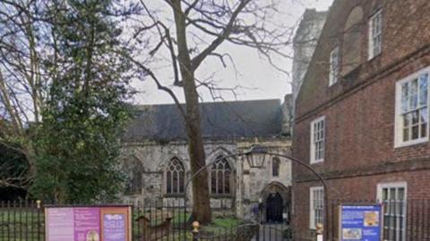The gated entrance to a church's grounds. The stone church building can be seen behind a large tree trunk. Some signs are placed either side of the arched gated entrance on the metal fence that sits around it.