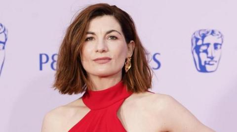 Jodie Whittaker looking at the camera. Her head and shoulders are visible and she is wearing a red dress. Behind her is a lilac background with pictures of the Bafta logo on it.