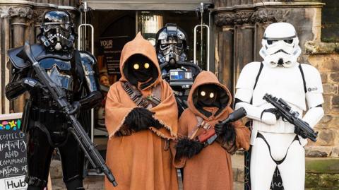 Three people dressed as Stormtroopers and two dressed as Jawas from Star Wars, pose for a picture outside of the entrance to a building.