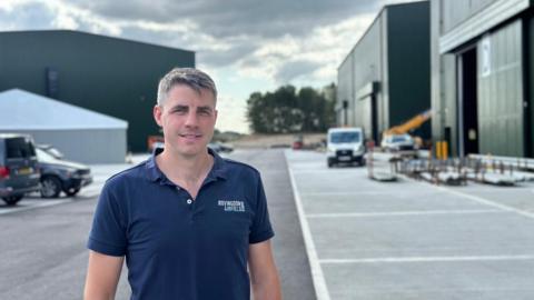 Harvey Mash, wearing a blue polo shirt with his company logo on his left breast stands outside film sound stages at Bovingdon Airfield Studios.