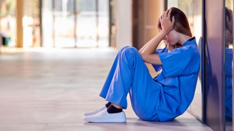A hospital worker looks burnt out. She is slumped to the floor and wearing blue overalls. 