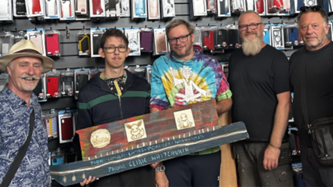 A group of five men stand in a phone shop, holding a miniature wooden narrowboat, which has been painted. On the side it says 'Phone Clinic Whitchurch'.