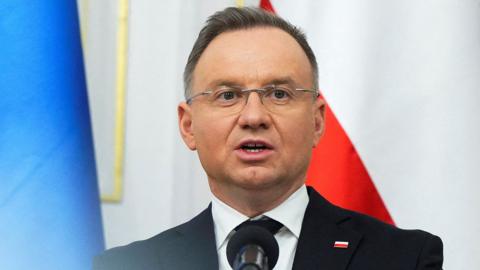 Poland's President Andrzej Duda looks away from camera while speaking into a microphone wearing a suit and tie with a Polish flag badge on his lapel, in front of the Polish flag with the Ukrainian flag just in view, in Warsaw on 15 January