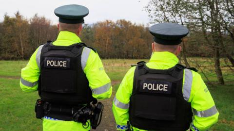 The back of two police officers. They are standing in a park with a row of trees in front of them. Both officers are wearing black caps, hi-vis coats and black vests reading "police" on the back. 