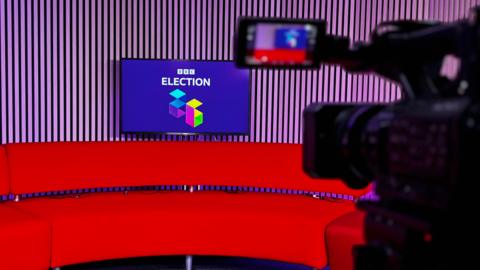 A red sofa in a broadcast studio, with wooden-panelled walls. There is a TV screen with a logo saying 'BBC Election 26' on it, on a purple background with white writing. In the foreground, a TV camera is visible.
