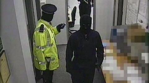 A gang member dressed as a police officer wearing a hi-vis jacket is standing with a gun in his hand. Two others, who are masked and wearing black are walking past him. There are notices on the wall in the depot.