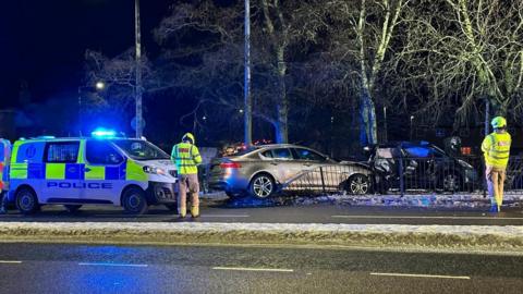 Crash scene in dark evening setting with police van with blue and yellow checks and blue lights, car crashed over fence, and another damaged car, and members of emergency services.