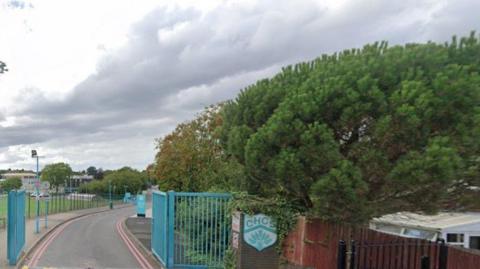 A generic photo showing open blue gates, with a sign to the right of them with the letters CHCS on it. A building and grass are on the far left and trees are on the right of the image.