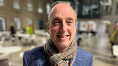 A middle-aged man smiles at the camera whilst wearing a tartan scarf and a rainbow lanyard with tables and other people obscured in the background.