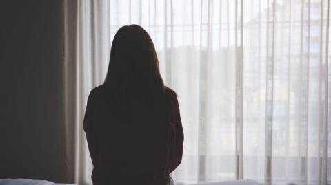 A woman, who is silhouetted, sits on a bed staring out of a window