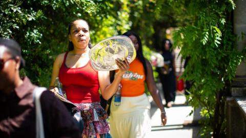 Woman fans herself with foil tray while walking under pergola