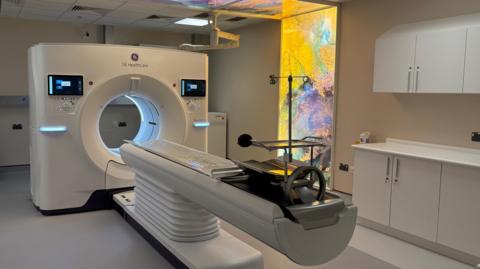 A CT scanner: a large square unit with a hole in the middle and a length of plastic, in the shape of a stretcher, sticking out into the room. The room is otherwise empty, aside from a series of kitchen-type units on the right-hand wall and a colourful light installation on the wall to the right of the scanner.