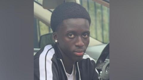Baye, a boy with short black hair, wearing a black and white striped tracksuit, looking at the camera. He is sitting in a car.