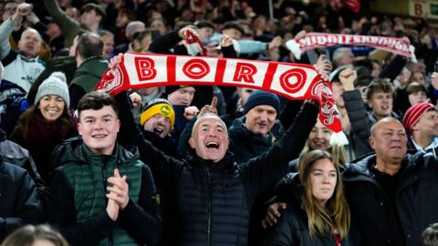 A Middlesbrough fan in the stands celebrates following the Sky Bet Championship match at Bramall Lane, Sheffield in february. He is holding a red and white scarf with Boro written across it and the club's crest. He is surrounded by other smiling and clapping fans. 