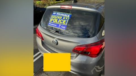 A silver car parked outside with a sticker on its rear window reading: "No Insurance: Seized by Police". There is a yellow square covering its licence plate. In the background you can see a green lawn. 