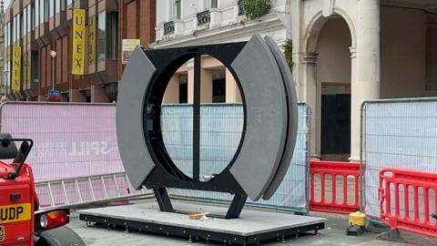 A black circle installation with gray side pieces was standing on concrete in the middle of the image. It was situated in the middle of a high street and had a silver barrier around it. 