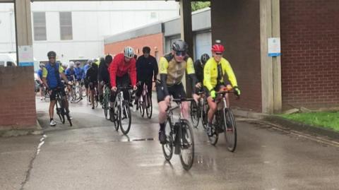 A large group of cyclists are setting off from Fulford Police station in York. They're all wearing helmets, and a large number of the group are also wearing sunglasses and in high visibility clothing.