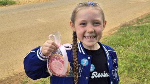 Woo, standing outside, holding up a biscuit in a bag, with a big smile. She has long hair, tied in a plait. She is standing by a grass area. She is wearing a black top and blue jacket.