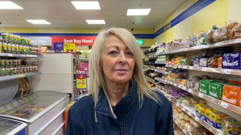 Nicola has long blonde hair and stands in the aisle of a supermarket.
