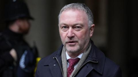 Communities secretary Steve Reed wearing a dark coloured coat, a check pattern scarf and a red tie. A police officer is stood in the background.