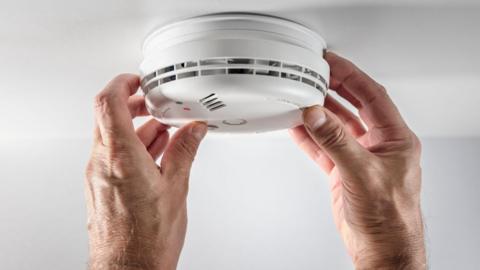 A pair of hands holding a fire alarm on a white ceiling