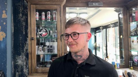 Kai Strevens stands in front of a cabinet containing hair products in the barber shop, looking to the right of the camera and smiling. He is wearing a black polo shirt, black framed glasses and has a large tatoo of an eye on his neck