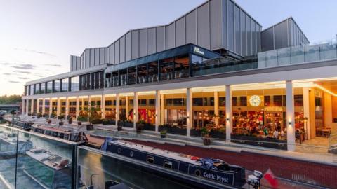 A new canal-side development features restaurants, a cinema and a rooftop bar. It is seen brightly lit at night with people gathered outside. Narrowboats are moored on either side of the canal below the development.