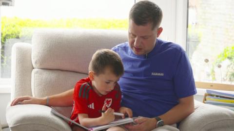 A man in a blue t-shirt, and a young boy with a red Wales football t-shirt are looking down at a coloring book. 