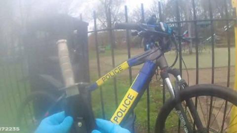 An image taken from body-worn camera footage showing an officer holding a sheathed knife in front of him. A police bike can be seen leaning on a park gate in front of him. 