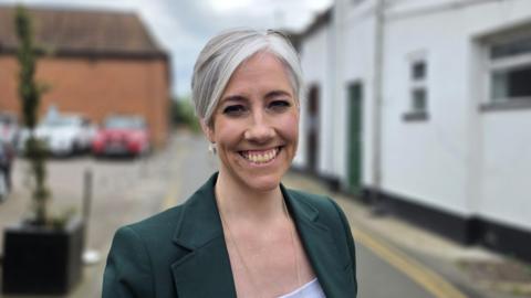 Daisy Cooper is standing in a street wearing a green jacket over a white top.