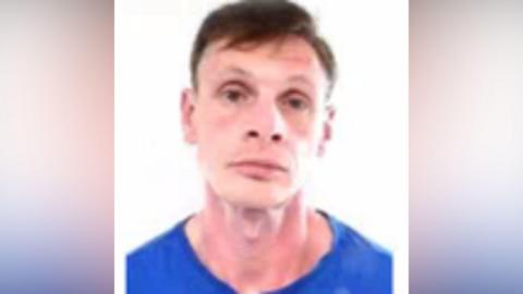 A mugshot of John Laidlaw. He has short brown hair with a side parting and brown eyes. He is wearing a blue top and looking at the camera with a serious expression.