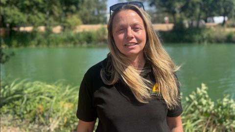 A woman with long blonde hair is standing in front of a fishing lake, wearing a black tee shirt and with sunglasses on her head.