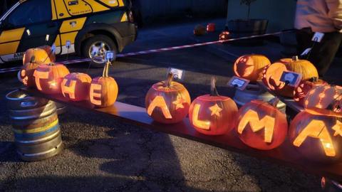 A row of pumpkins each carved with a letter to read THE ALMA. They are on benches on tarmac with a car in the background.