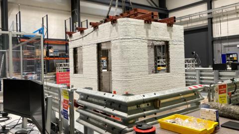 A white 3D printed structure in the shape of a building has been erected in a laboratory. The structure is being held down by steel. There are warning signs and a yellow tray full of protective eyewear.