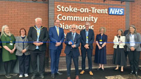 A man is cutting a blue ribbon with a line of people around him. Behind him is the wall of a building and a white sign which can be partly read and says "Stoke on Trent NHS Community Diagnostic". 